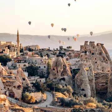 Village of cave dwellings carved into cone-shaped rocks in Göreme, with a minaret and hot-air balloons in a hazy dawn sky.