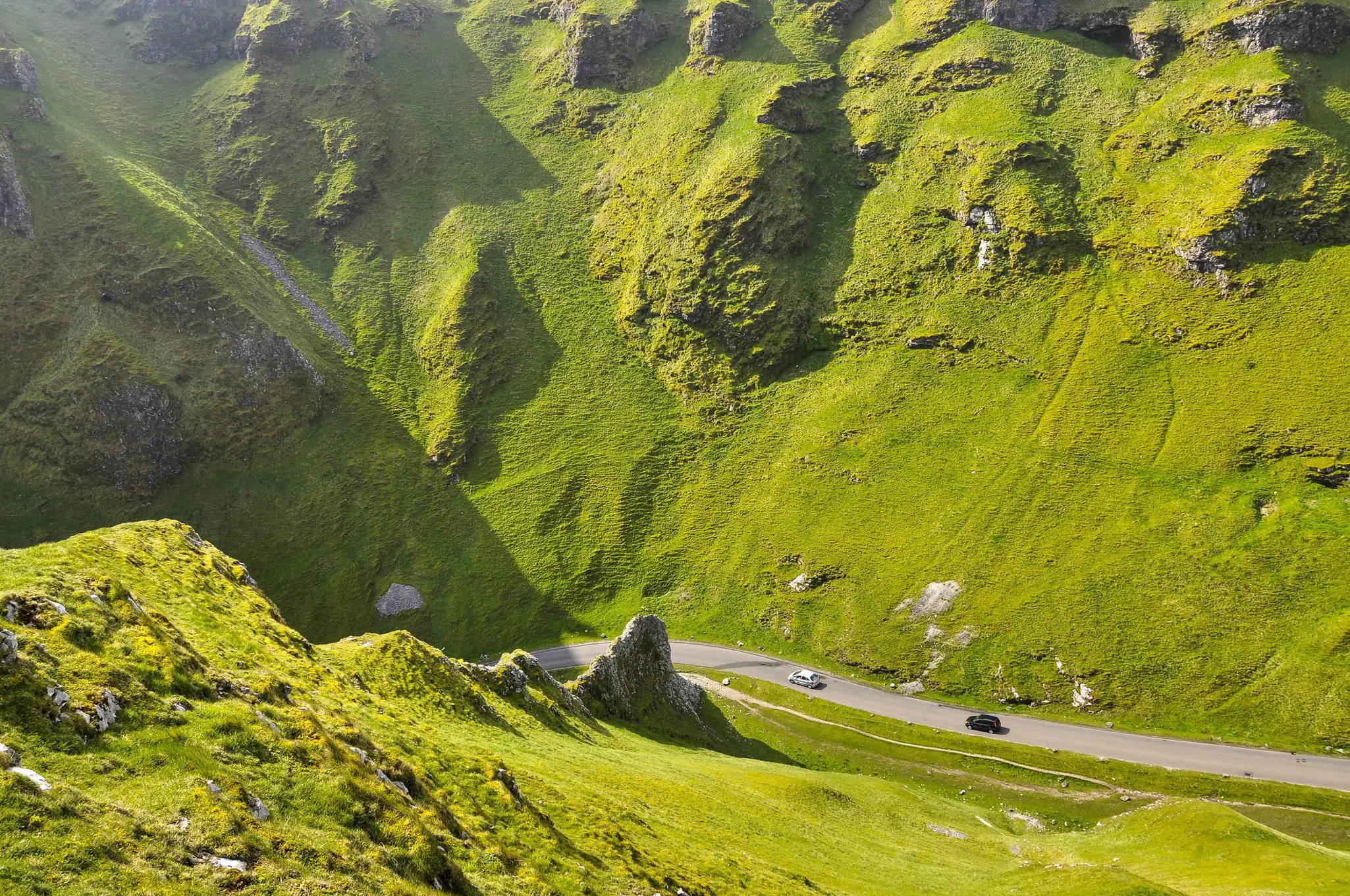 A wide shot of a green cliffs plunging toward a road with two cars.