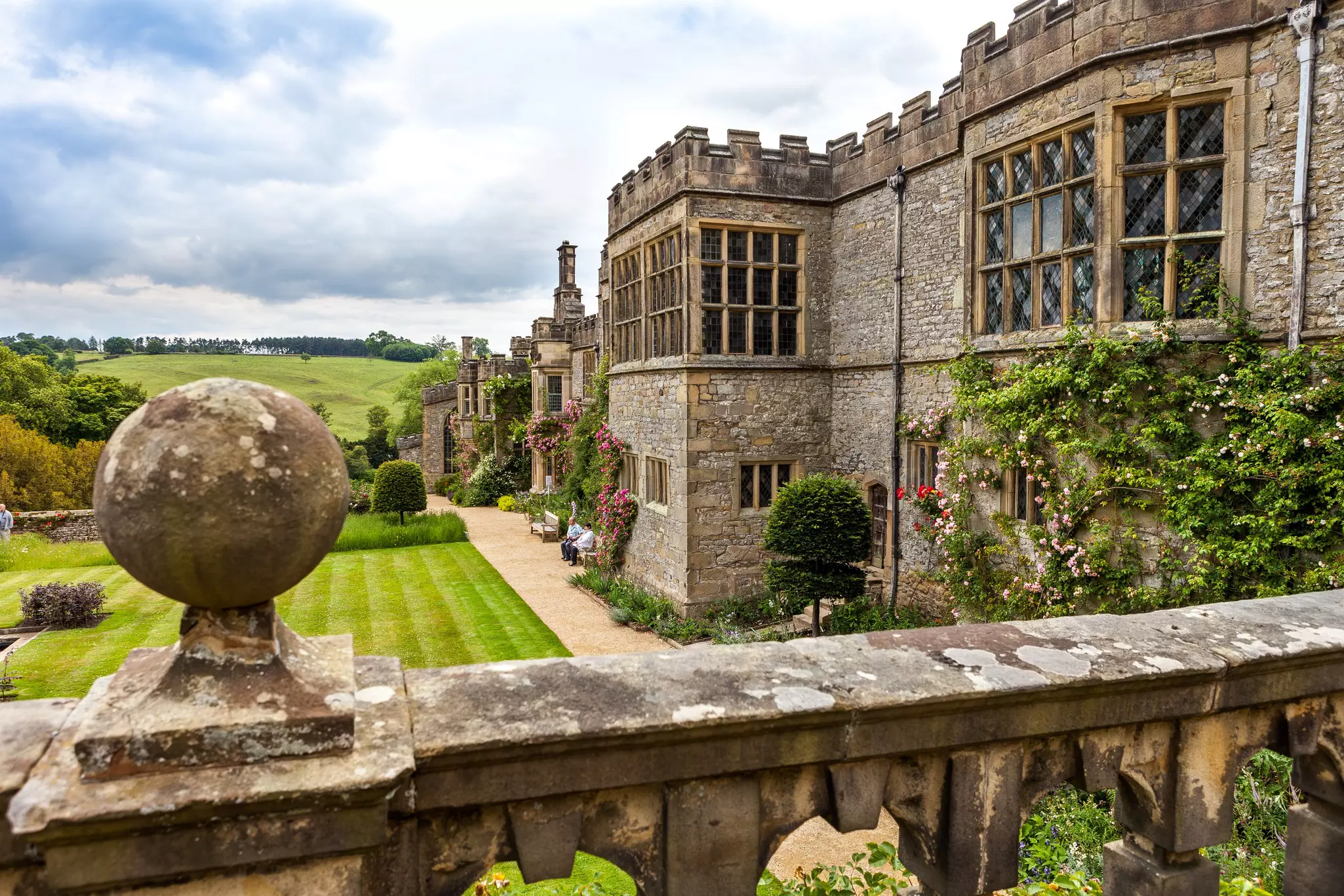 The exterior of a Tudor stately home, with tourists relaxing in its manicured gardens.