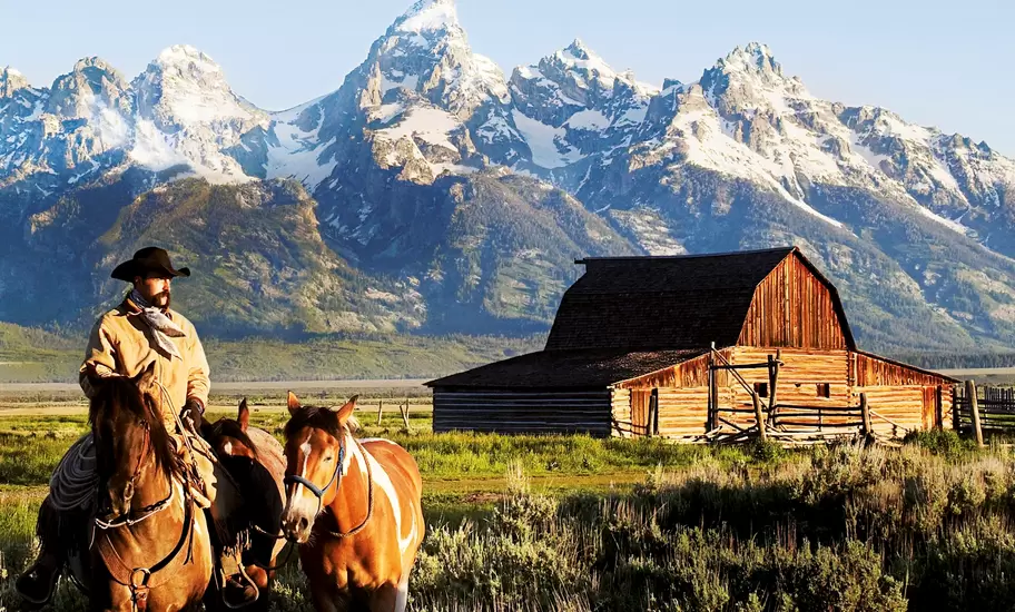 A cowboy on horseback leads another horse in front of a historic barn and the snow-capped Grand Tetons in Mormon Row