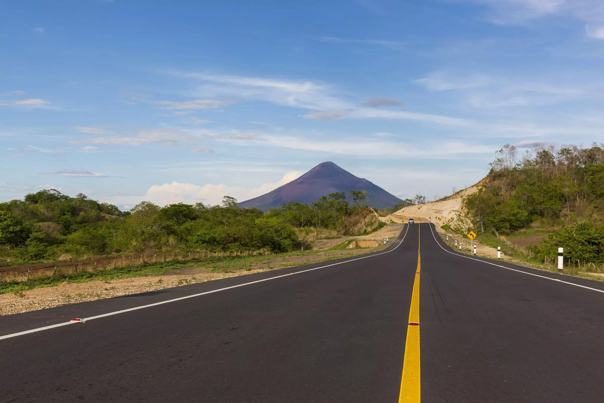 A long, smooth, asphalt road leads through countryside towards a pointed volcano.