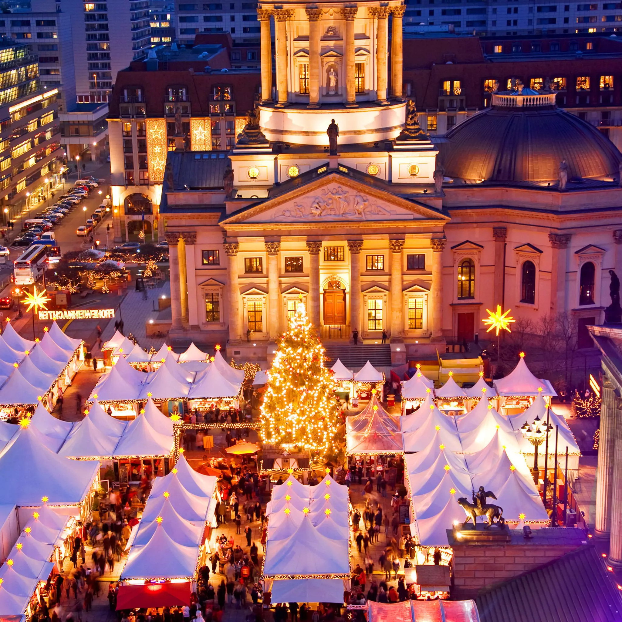 People shop at a line of tent-covered stalls at night, seen from above, surrounded by white Christmas lights and a lit Christmas tree, with city buildings in the distance.