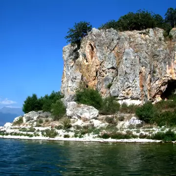 A pale rock island rising sheer from dark green water, topped with trees and scrub, a cave mouth in its face and distant mountains across the lake.