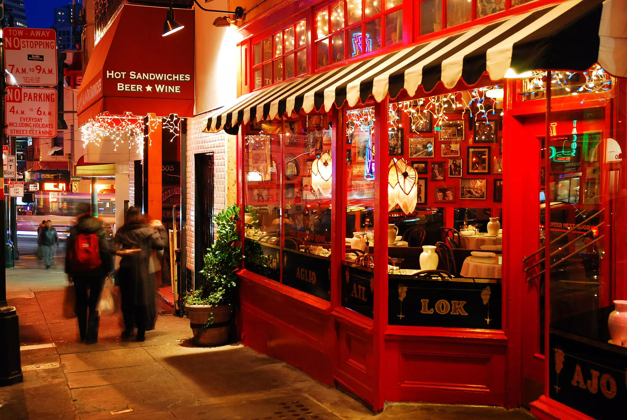 Nighttime street scene with red bistros to the right and out-of-focus people in the distance.
