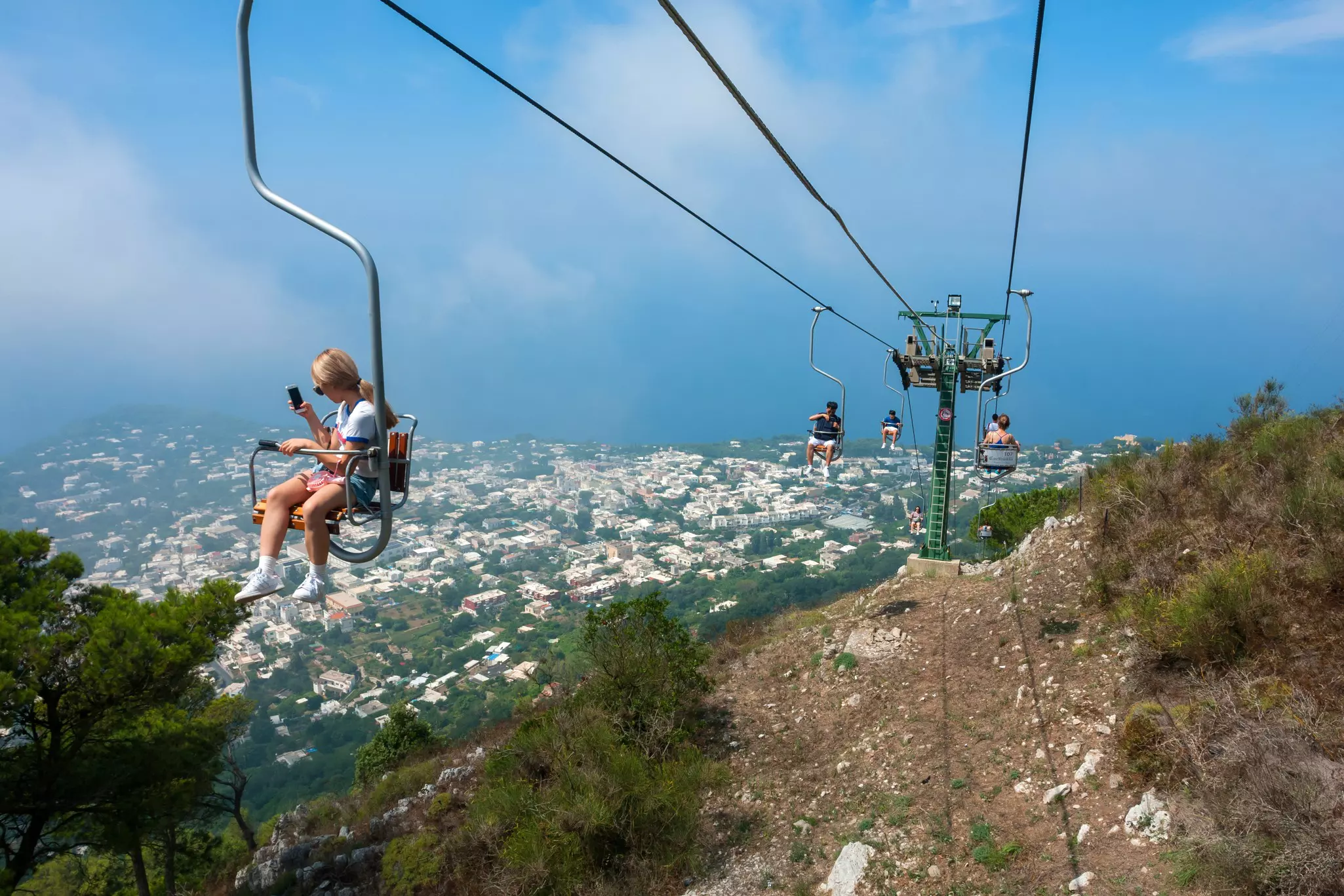 Tourists riding up the mountain chairlift on the island of Capri, Italy, with a view of houses and the coast behind.