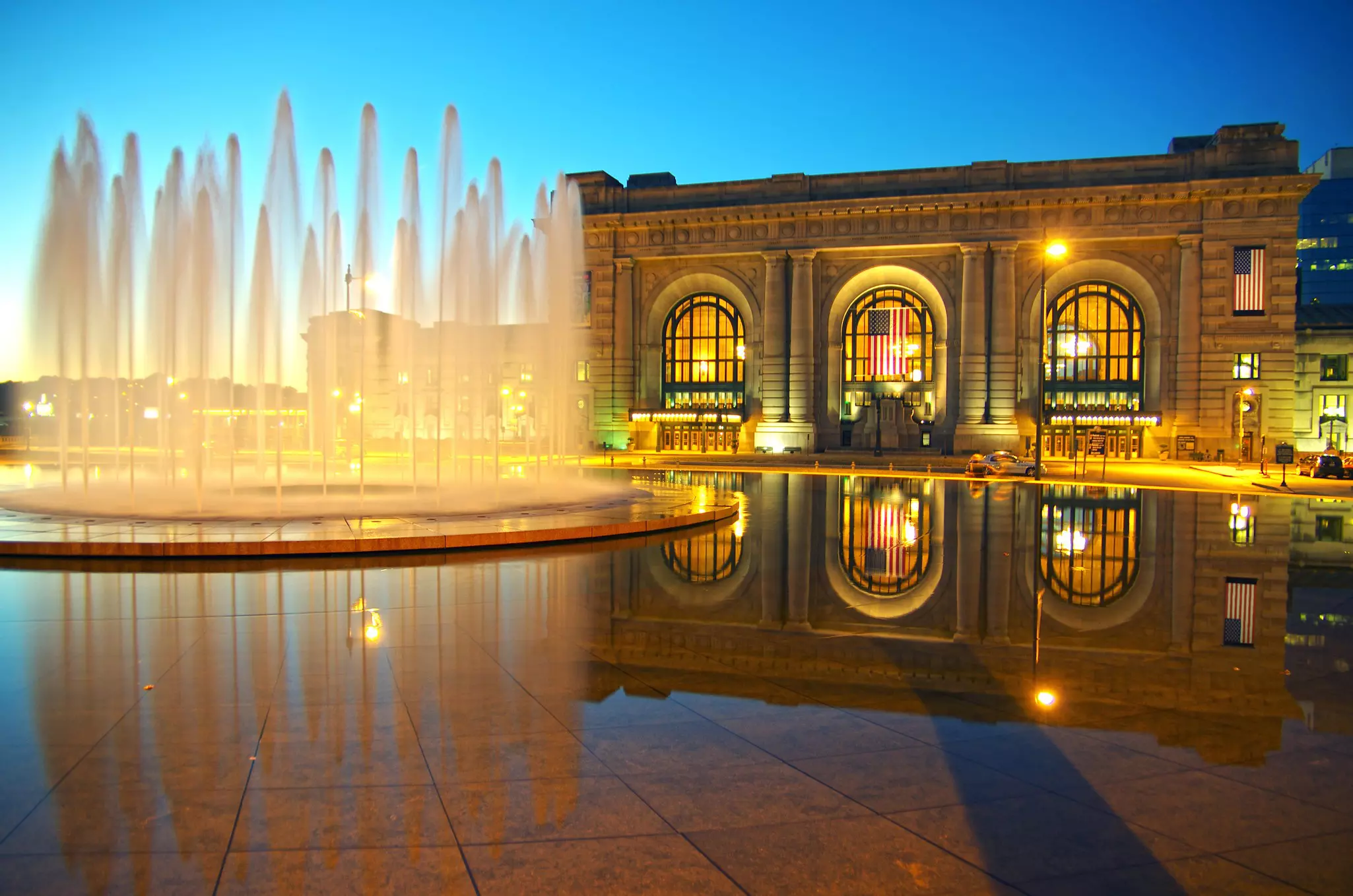 Streams of water is illuminated by blue light spray from a stone fountain. In front of the fountain is a man riding a large horse.