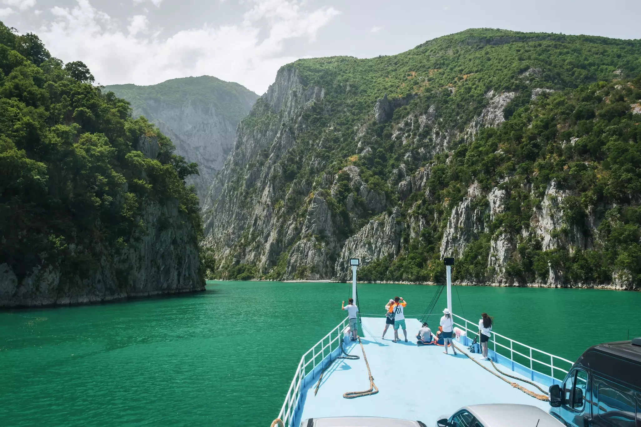 People on the deck of a ferry on a wide green lake with sheer cliffs either side.