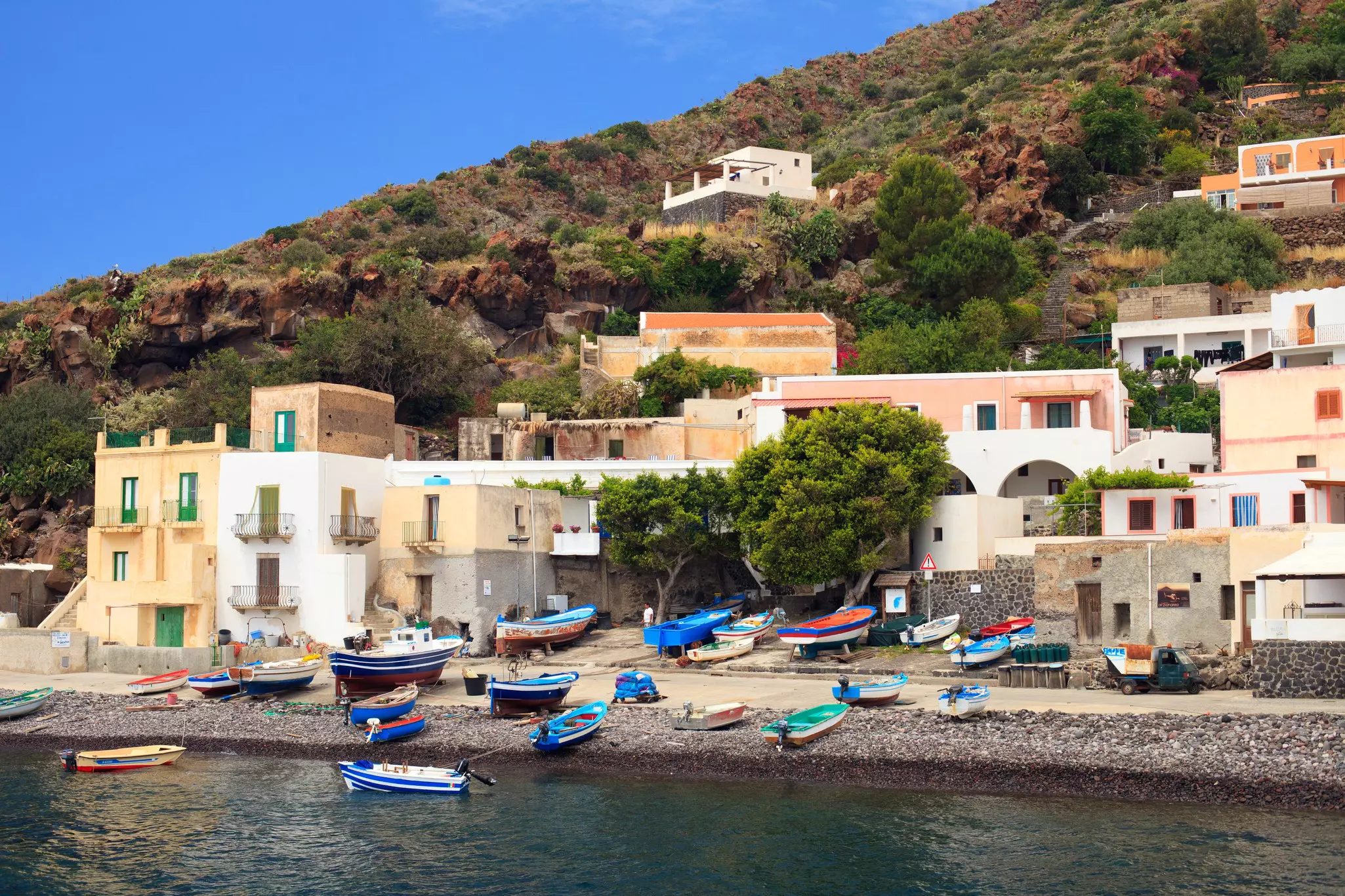 Colorful boats pulled up on a rocky shore with flat-roofed houses along the roadway on a sunny day.