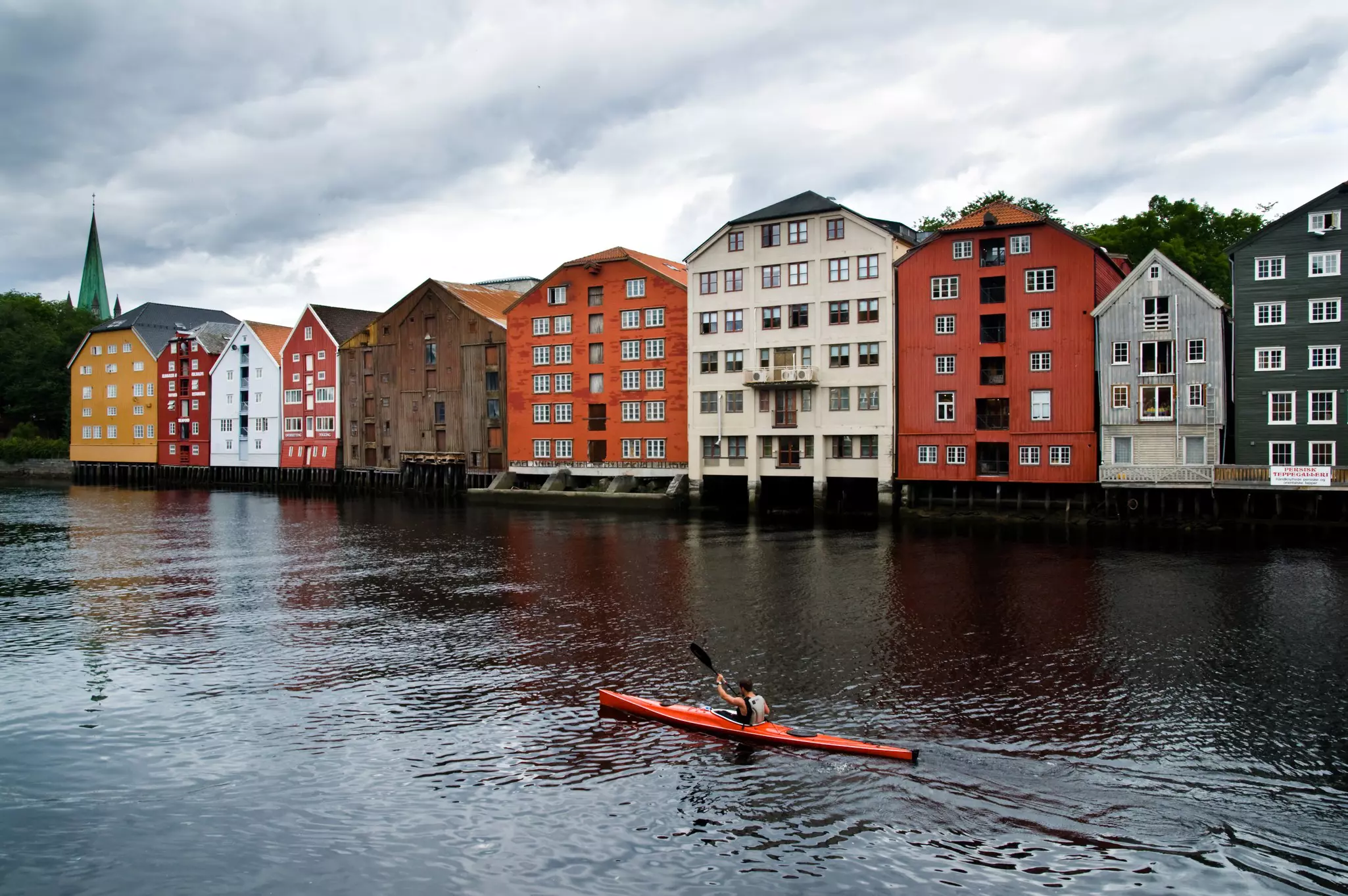 A person in an orange kayak paddles in a river in Trondheim, Norway. Brightly painted houses line the riverbank.