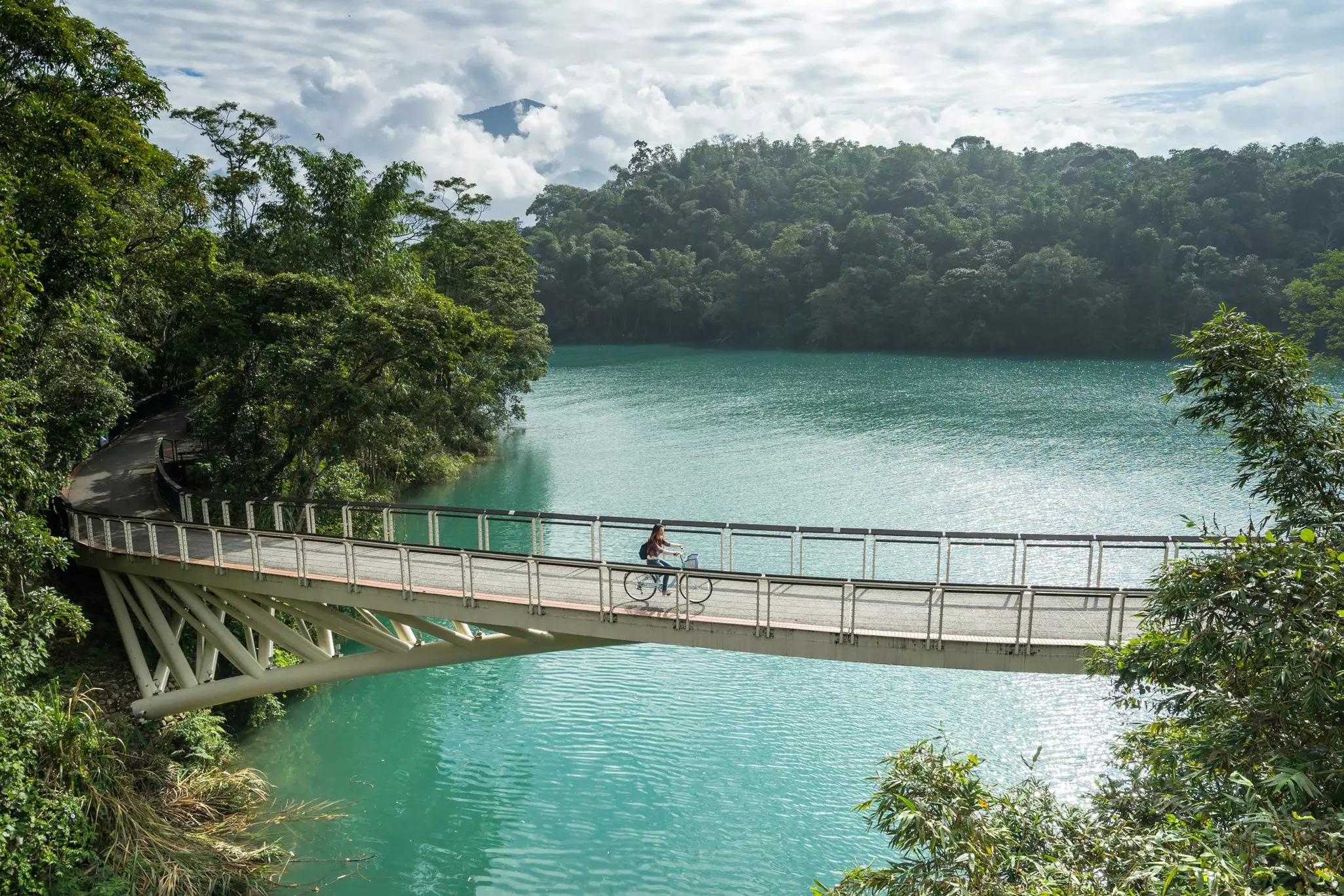 A solo female cyclist rides a bije over a bridge on a trail around a lake.