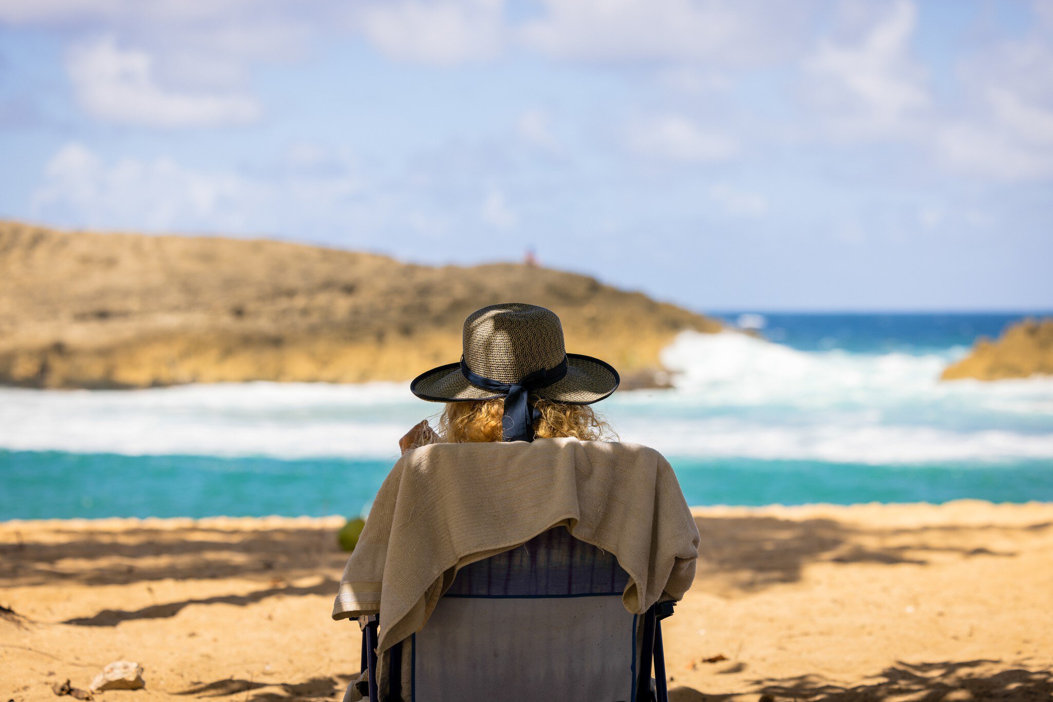 The back view of a woman sitting in a chair on a sandy beach in the shade with a sun hat on and towel draper over the chair's back. The blurred ocean can be seen in the close distance on a sunny day.