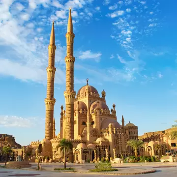 Al Sahaba Mosque in Sharm El Sheikh with two tall ornate sandstone minarets, domes and palm trees under a blue sky