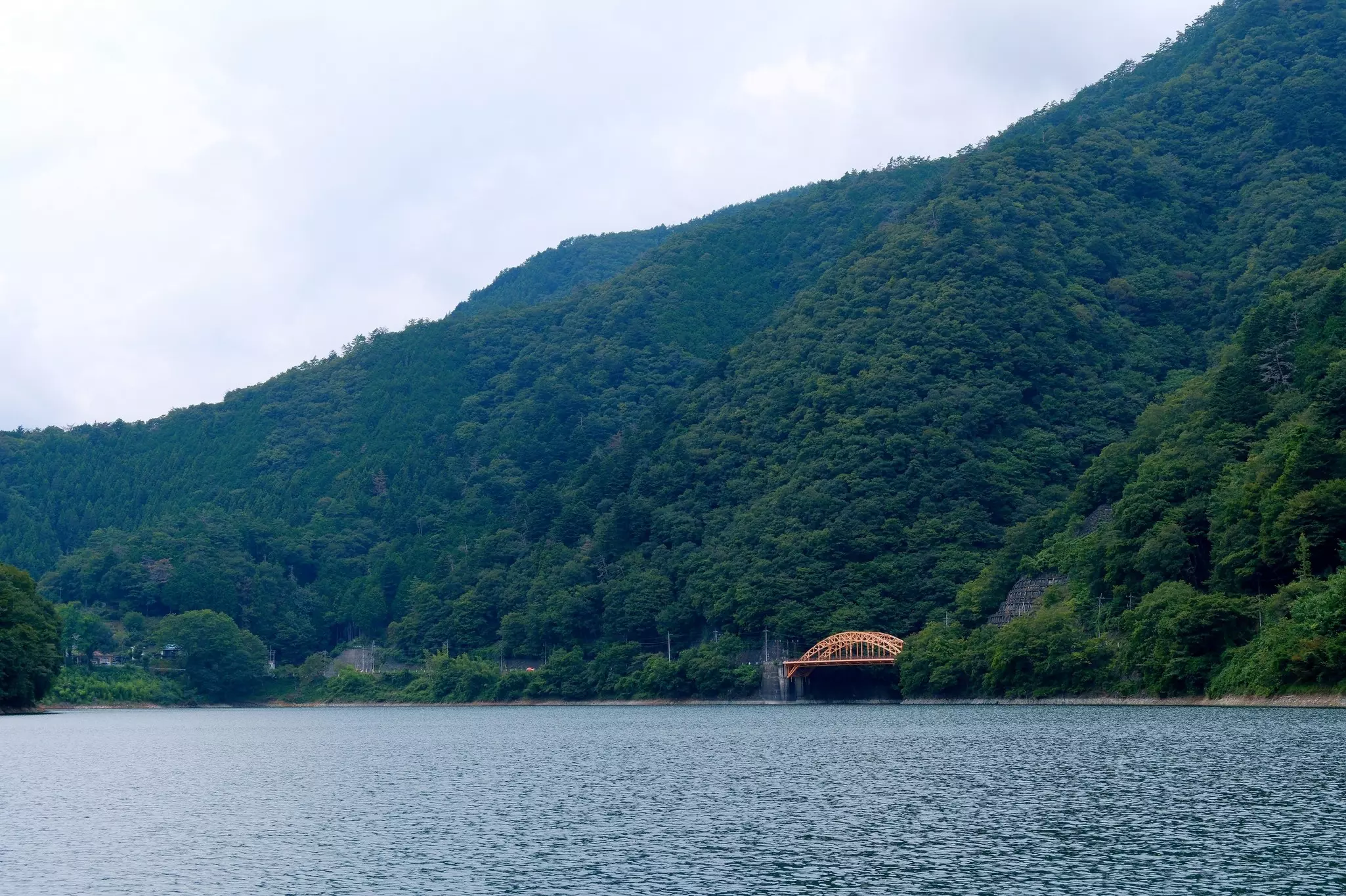Lake view with an arched bridge and forested mountain in the distance on an overcast day.