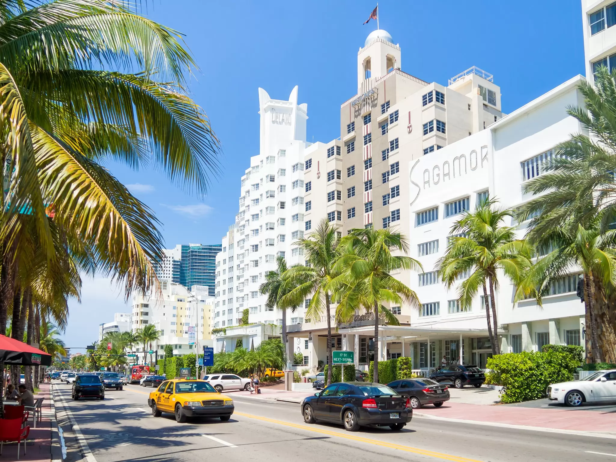 A row of art-deco hotels on a busy street lined with palm trees.