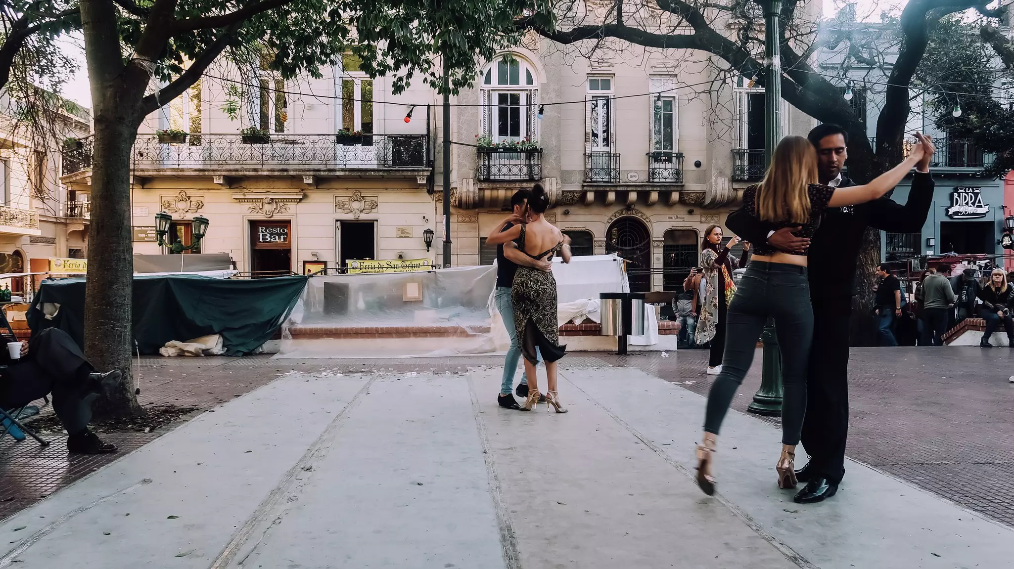 Couples dance the tango in a city square. Historic buildings line the plaza.