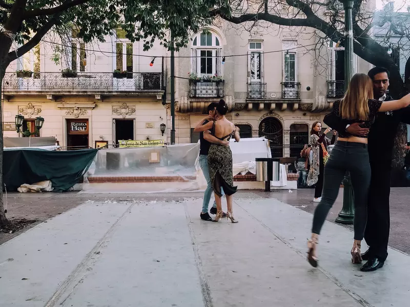 Couples dance the tango in a city square. Historic buildings line the plaza.