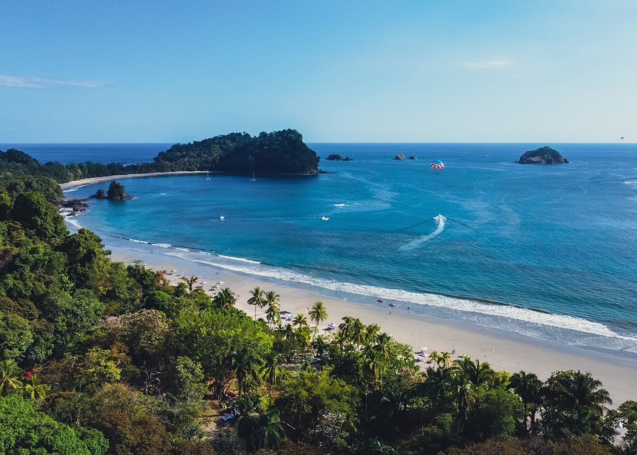 Aerial view of forest in foreground leading to curved sandy beach and the ocean and rocky outcroppings beyond. A few boats are in the sea, as well as a parasailer in the air.