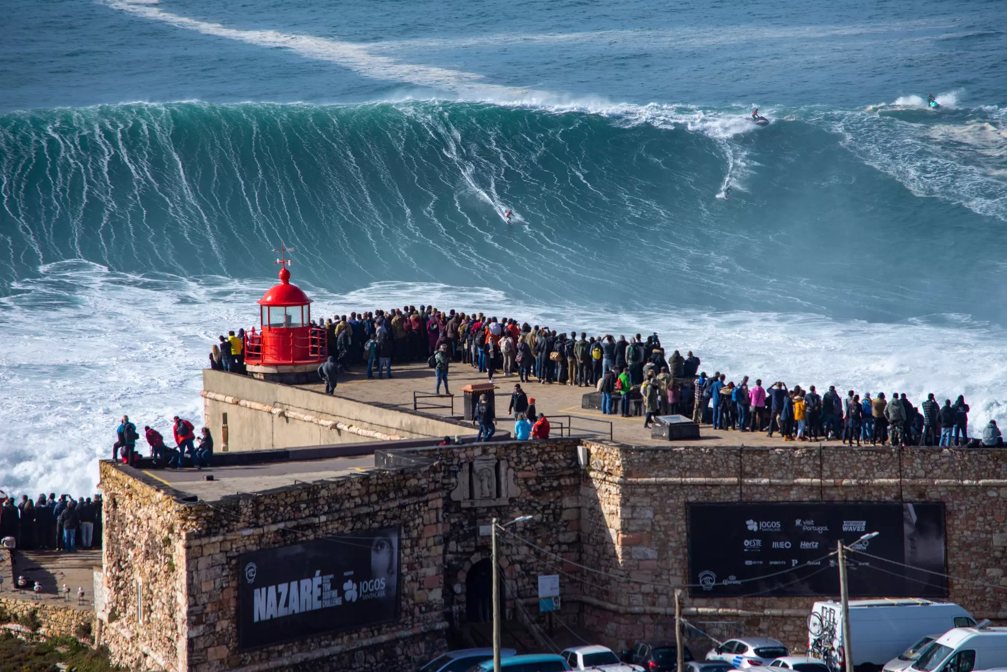 Spectators gather at a vantage point near a lighthouse to watch surfers tackle massive waves.