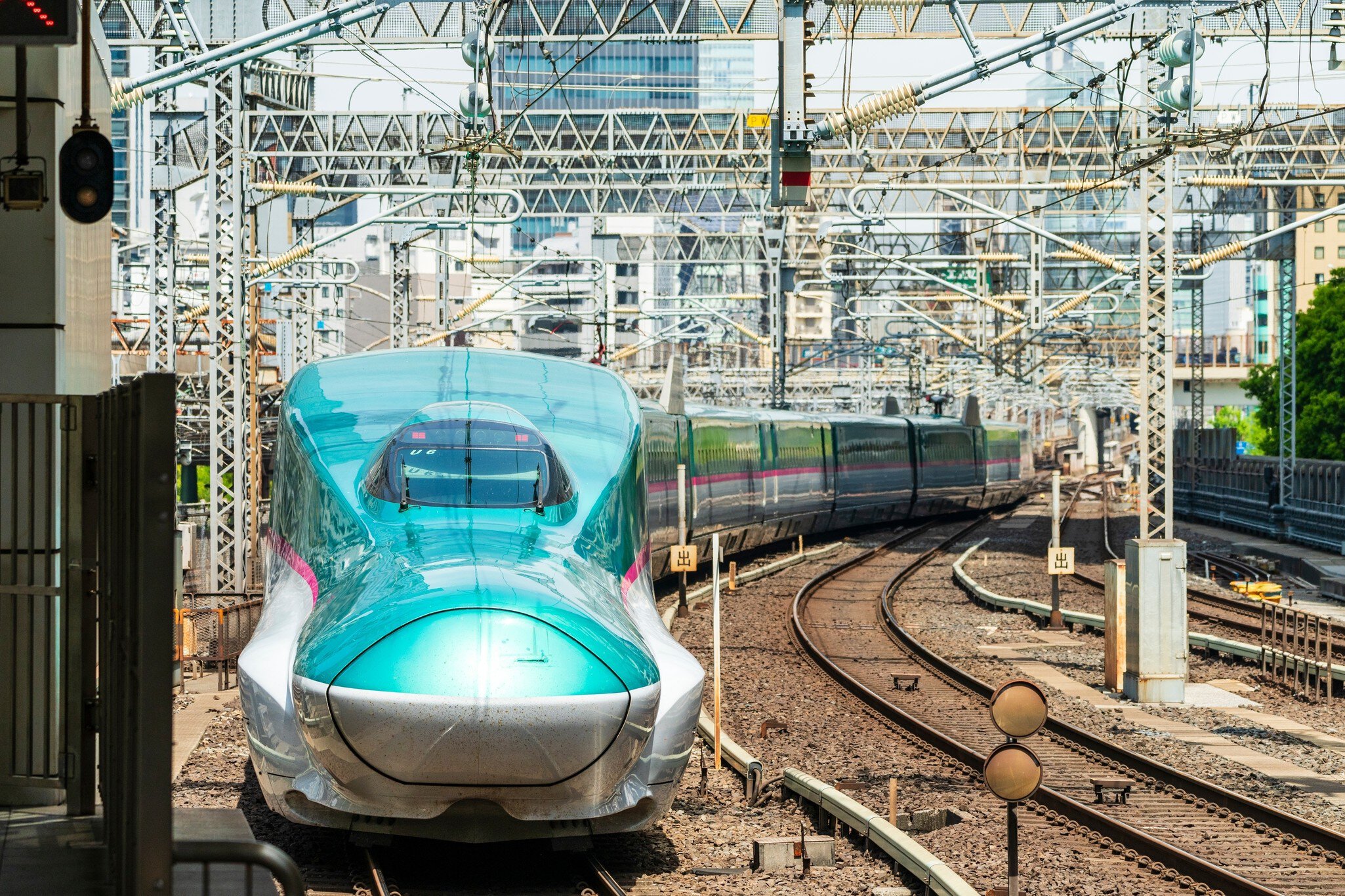 A teal-colored train with a bullet-shaped front carriage travels on tracks out of a city.