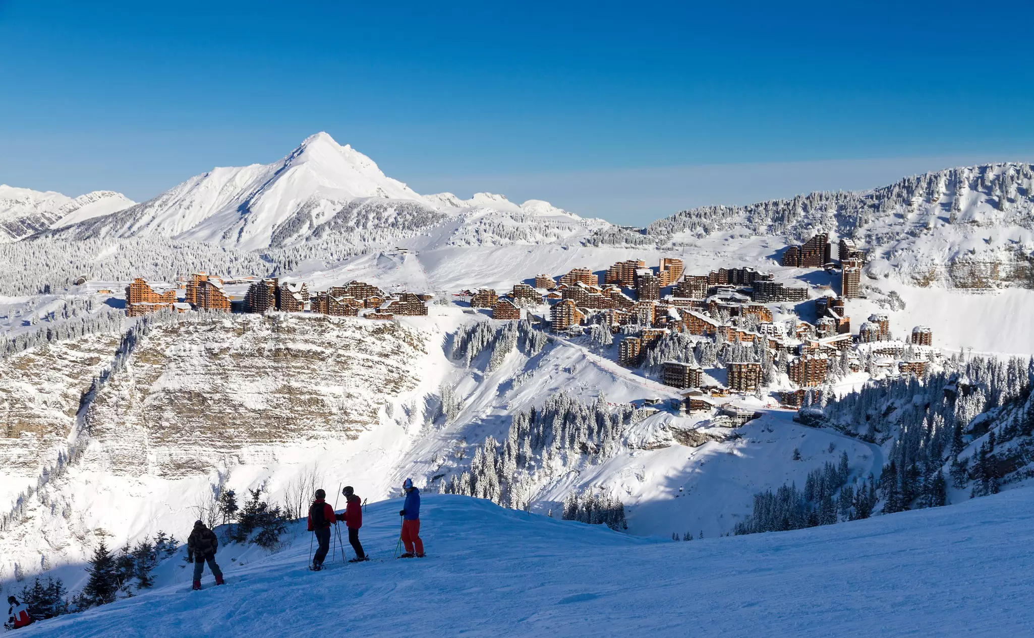 Skiers on a slope. Behind them across the snowy valley a resort is built into the side of a mountain