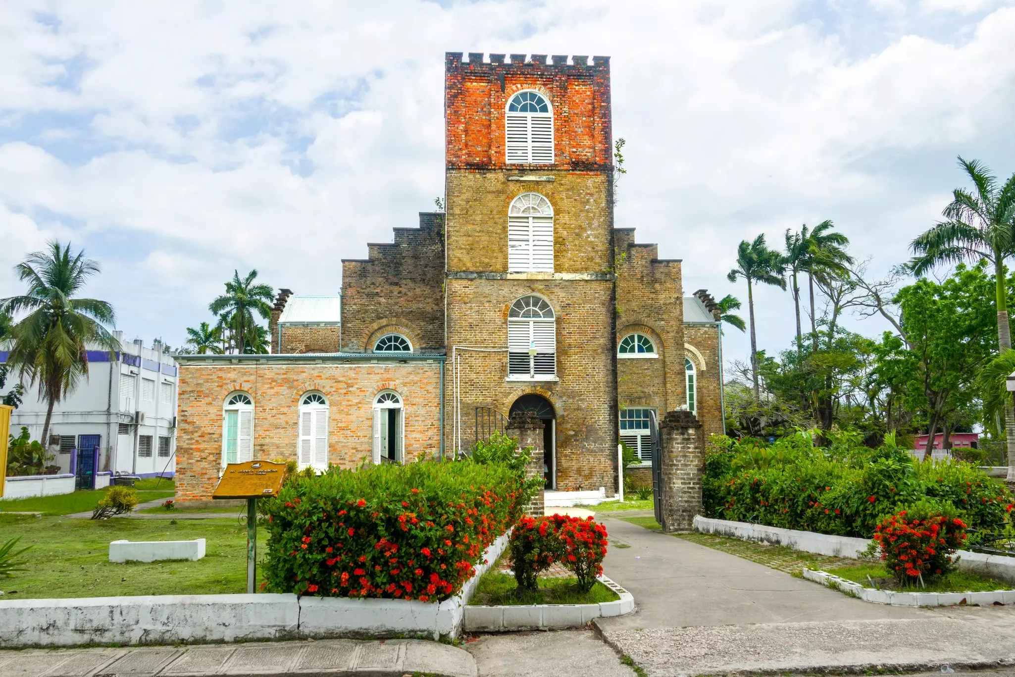 Exterior of a brick churck with palm trees in the background and red-flowered bushes in front on a bright, cloudy day.