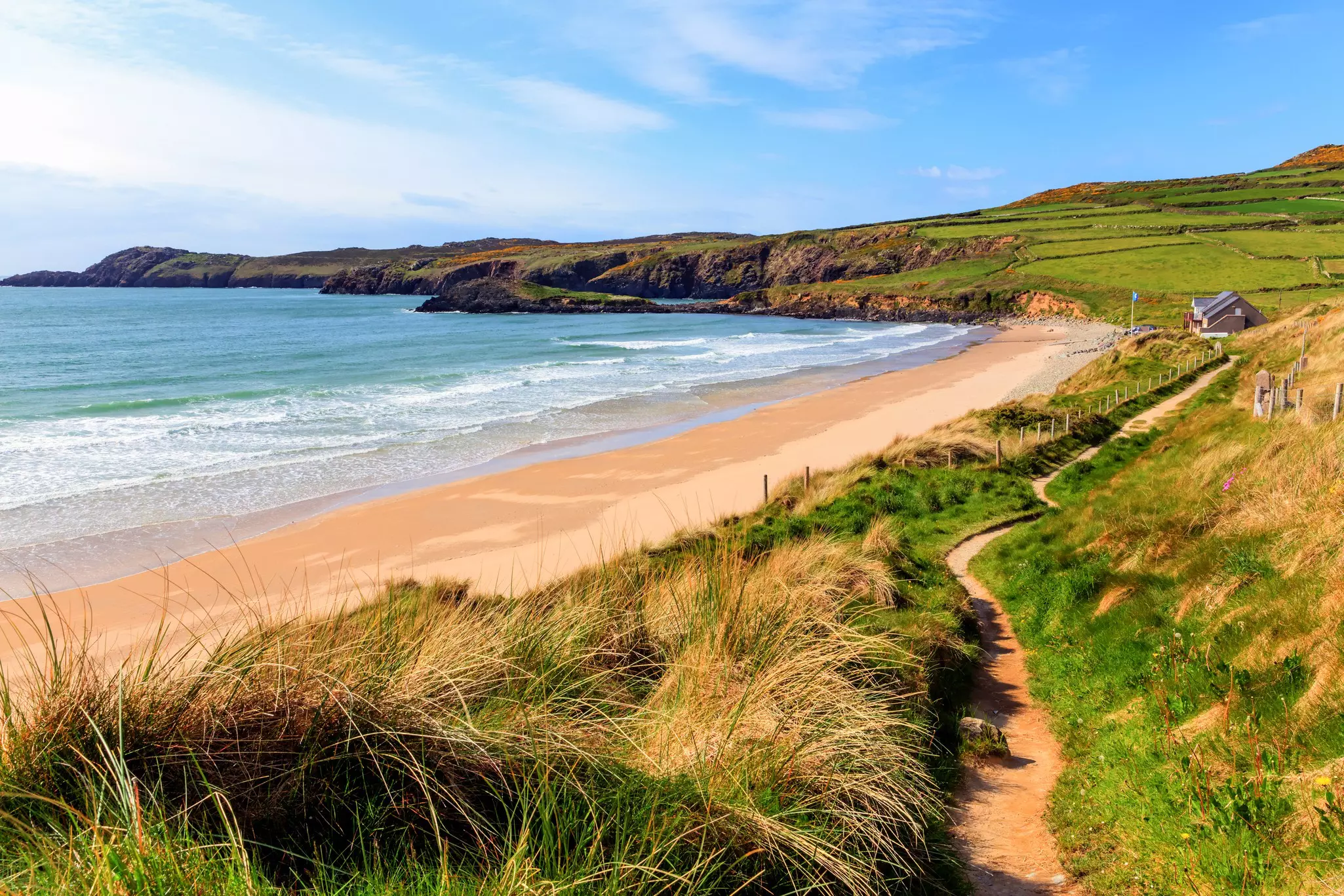 A golden curve of sand backed by grass-topped cliffs on a sunny day. A path follows the top of the cliff.
