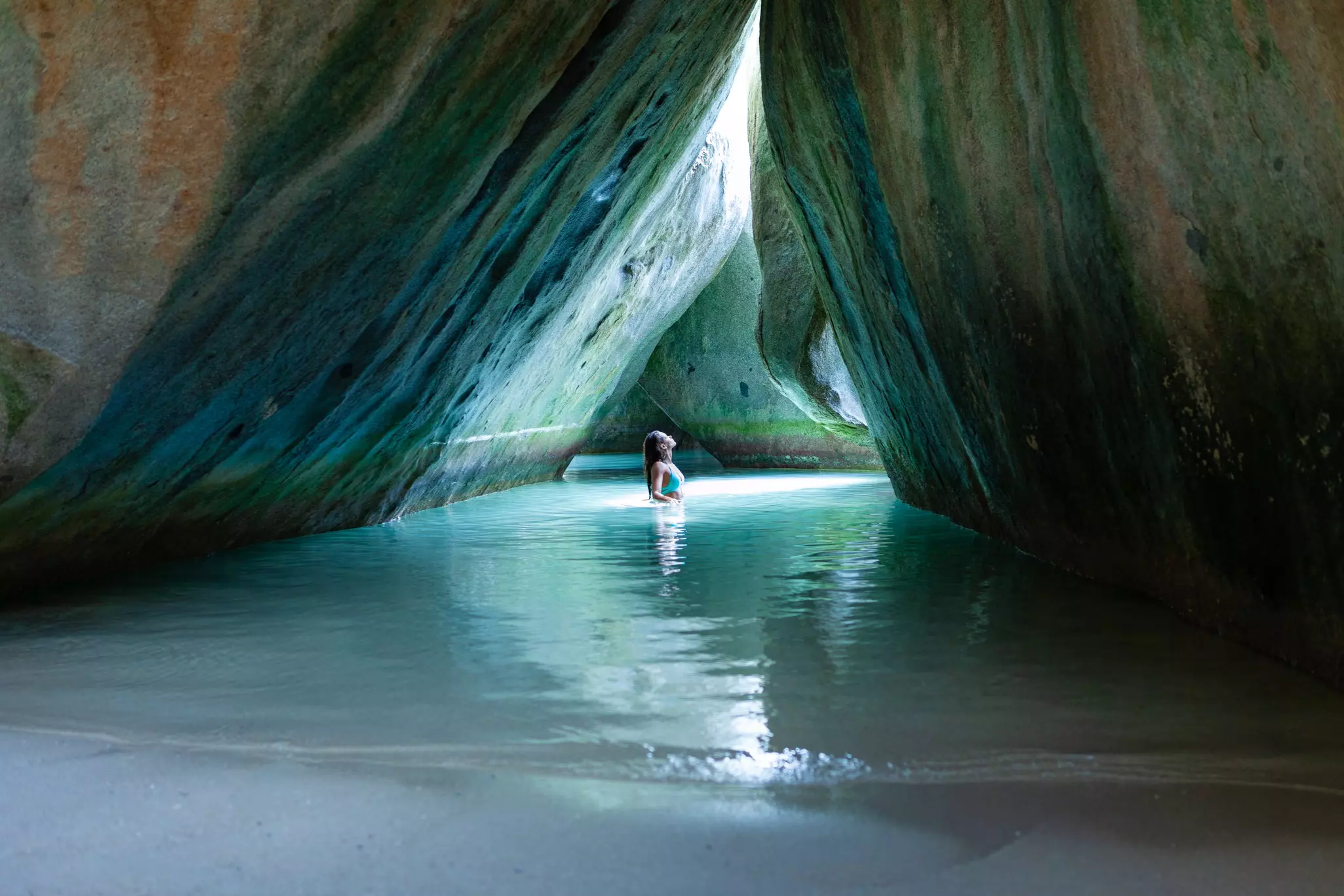 A woman is waist-deep in water in a cave by a beach, looking up a beam of light that is hitting her body.