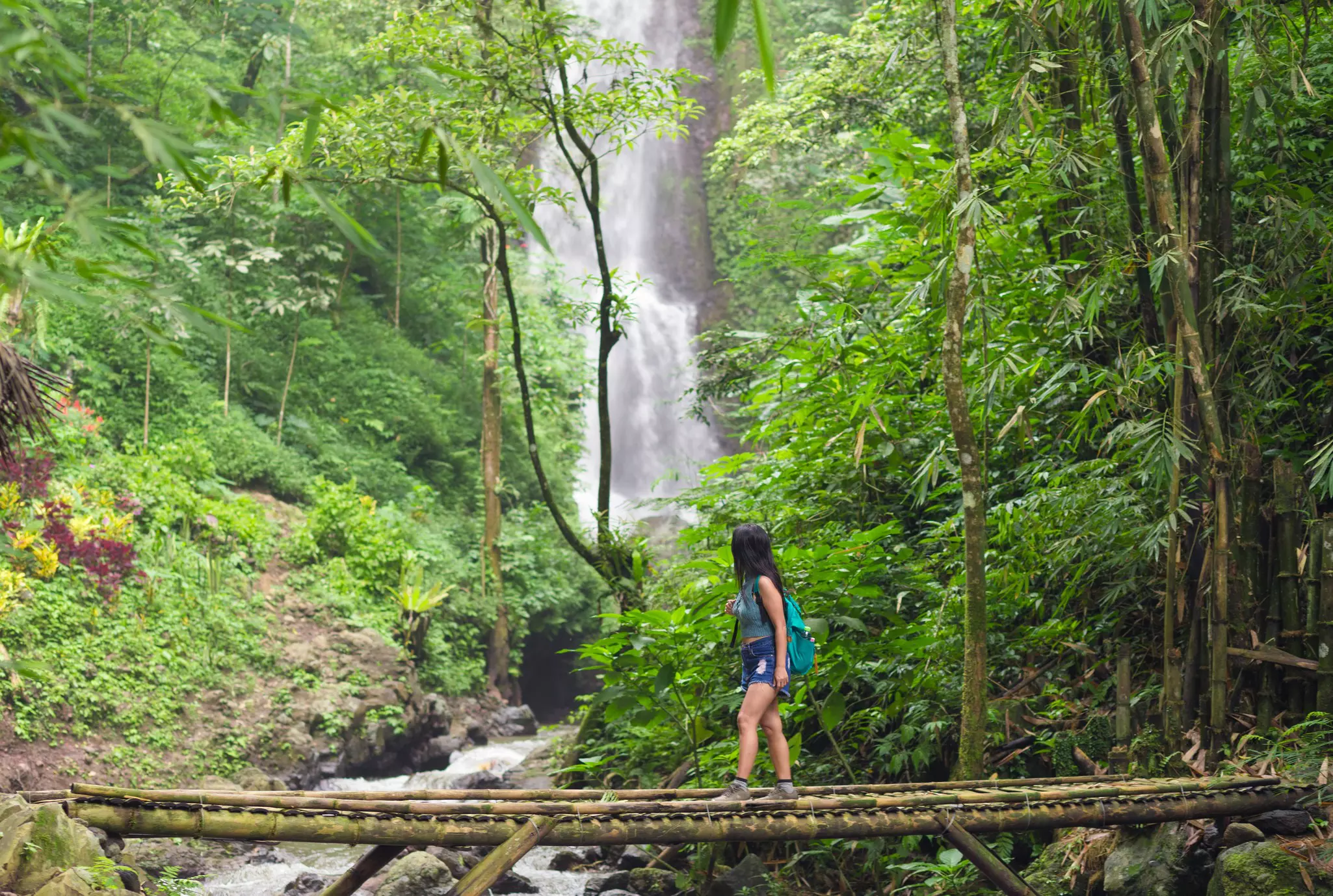 A female hiker crosses a bridge in a jungle, turning her head toward a waterfall in the distance