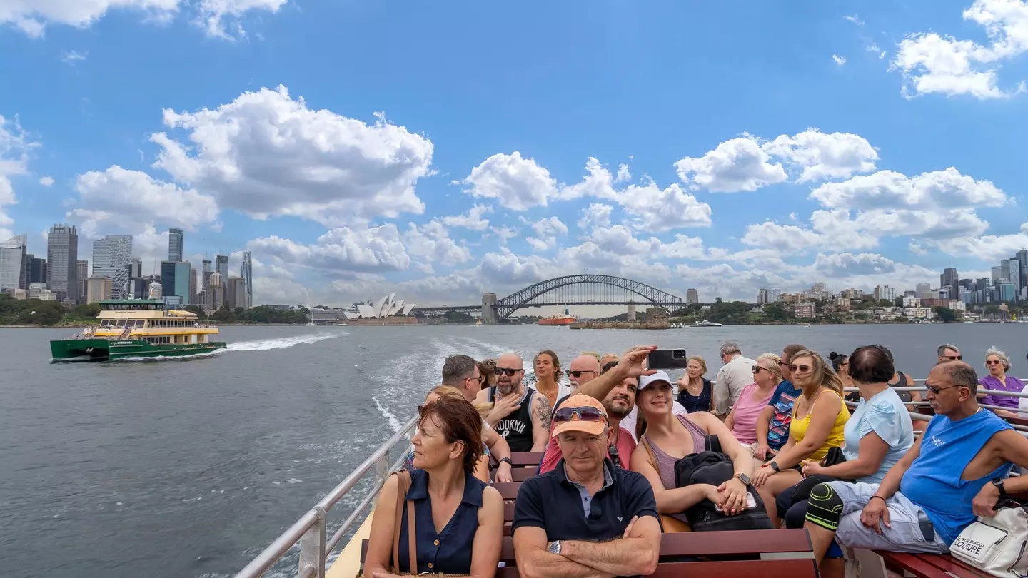 People sit on an exterior deck on a ferry passing through the harbor of a busy city with a big bridge and skyscrapers in the distance.