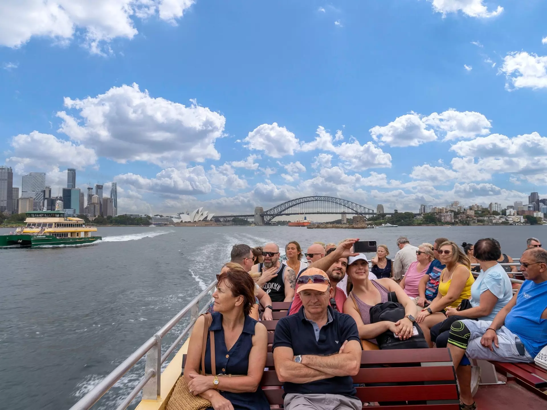 People sit on an exterior deck on a ferry passing through the harbor of a busy city with a big bridge and skyscrapers in the distance.