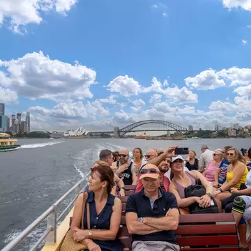 People sit on an exterior deck on a ferry passing through the harbor of a busy city with a big bridge and skyscrapers in the distance.