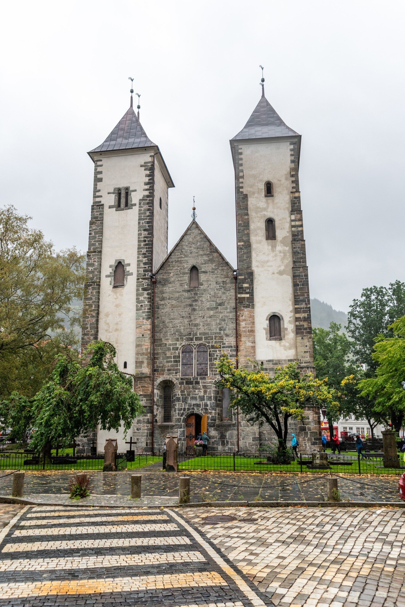Romanesque stone church with two white-plastered towers and grey pyramid roofs above a cobbled street, overcast sky