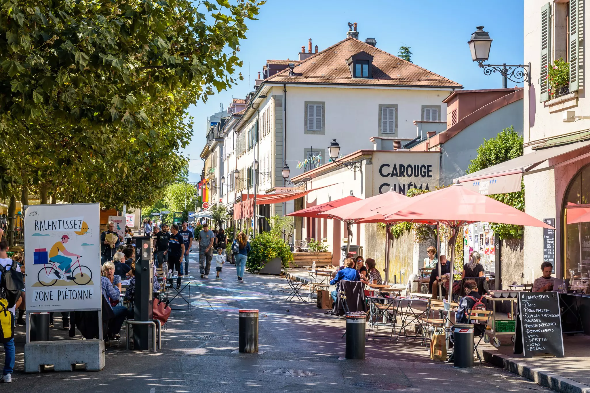 A street in a city is closed to cars, lined with outdoor tables and filled with pedestrians who walk past shade-giving trees.