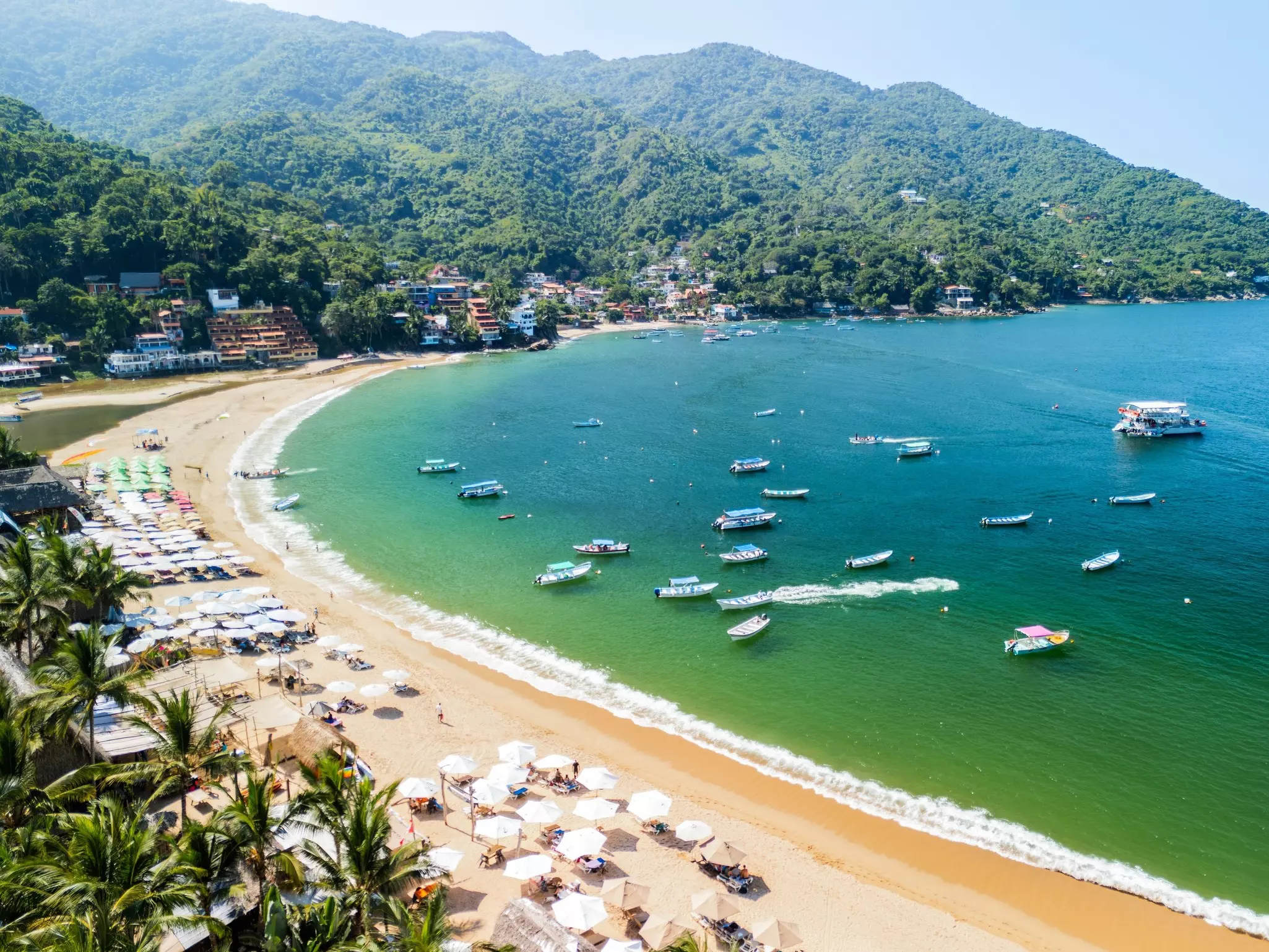 An overview of wide beach by a bay with numerous umbrellas in the sand and boats in the water.