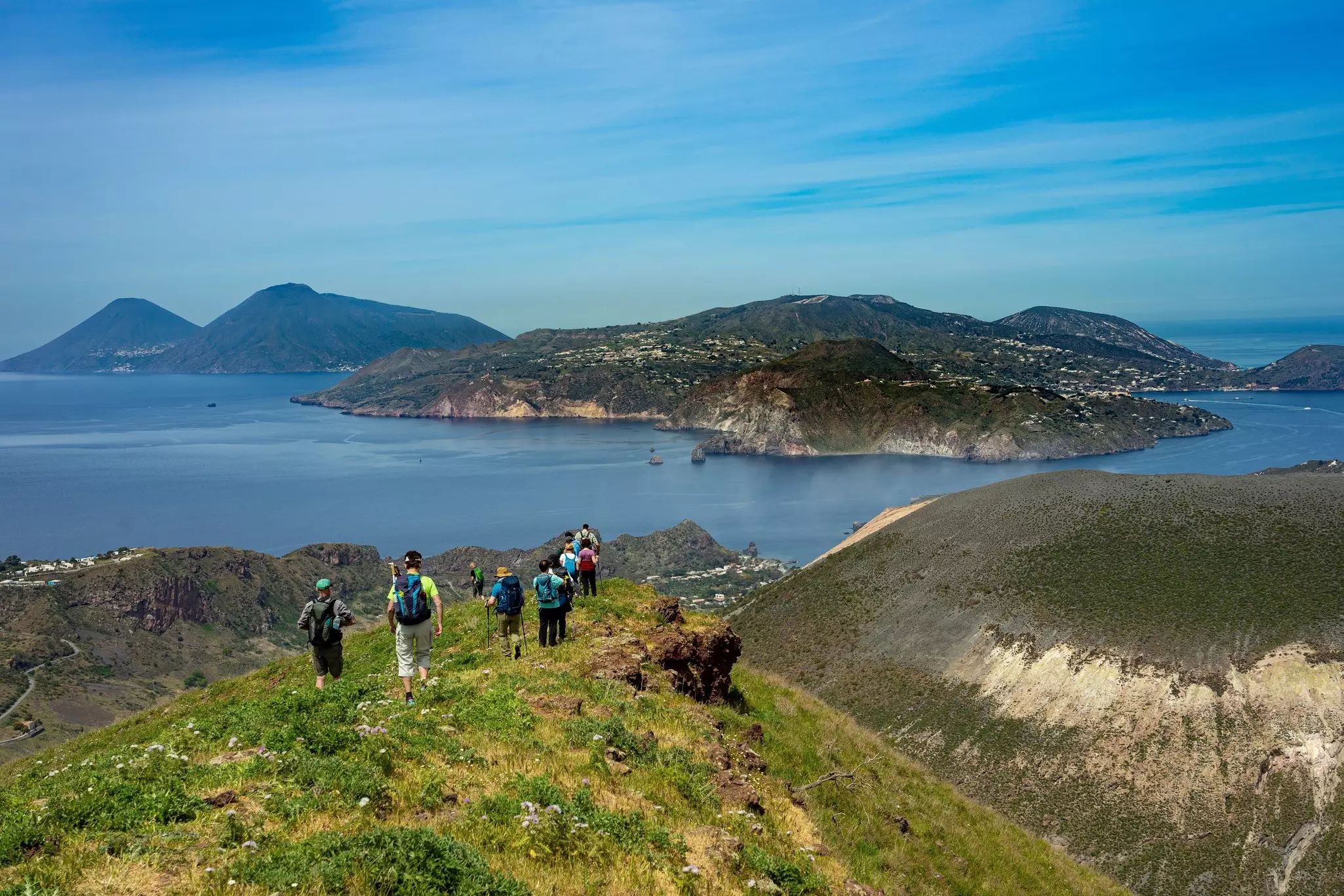 A group of hikers walk through a mountain meadow heading down towards the coastline.