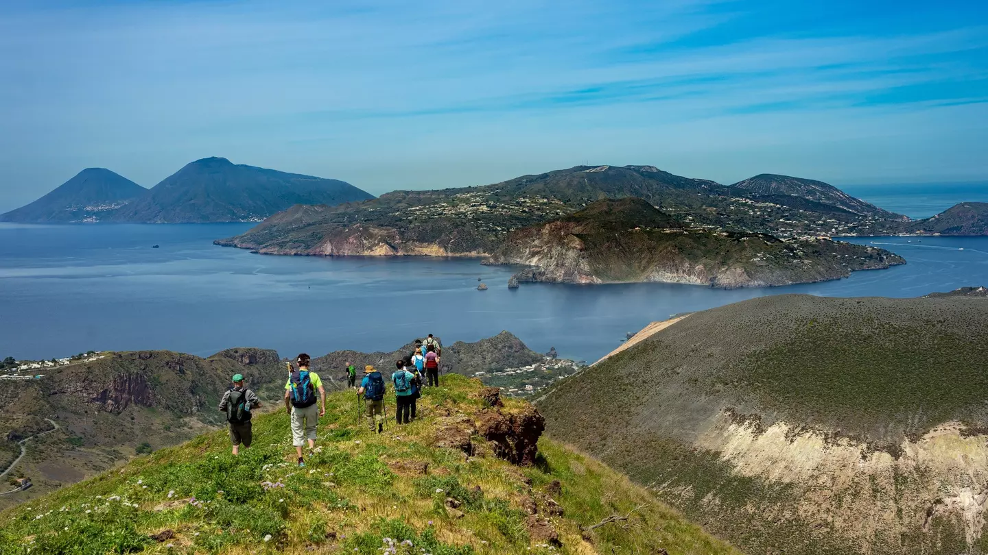 A group of hikers walk through a mountain meadow heading down towards the coastline.