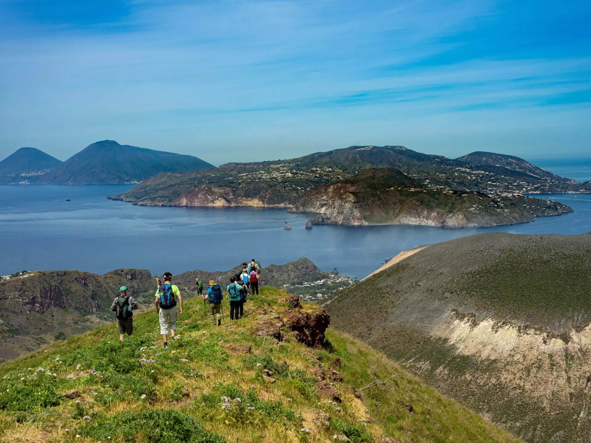 A group of hikers walk through a mountain meadow heading down towards the coastline.