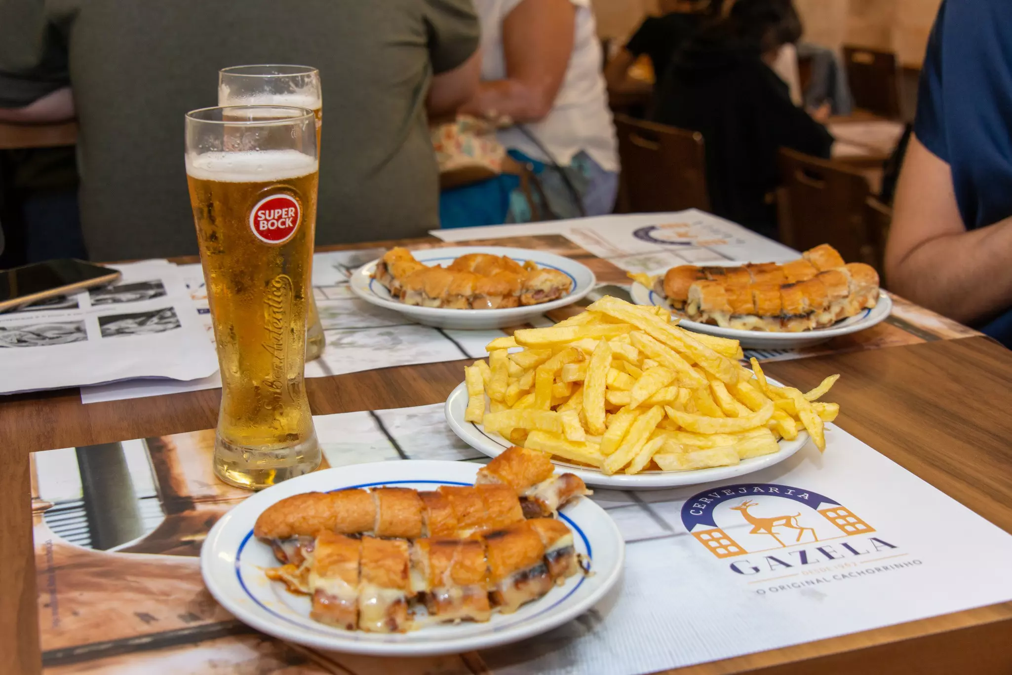 Three plates of a small hot dog made of pork sausage with melted cheese and stuffed in a crispy baguette that is brushed with butter and a spicy sauce, surround one plate piled with french fries on a wooden table beside two tall glasses of beer