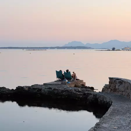 People sit in lawn chairs out on a rocky outcrop, looking out at the sea and hills in the background. 