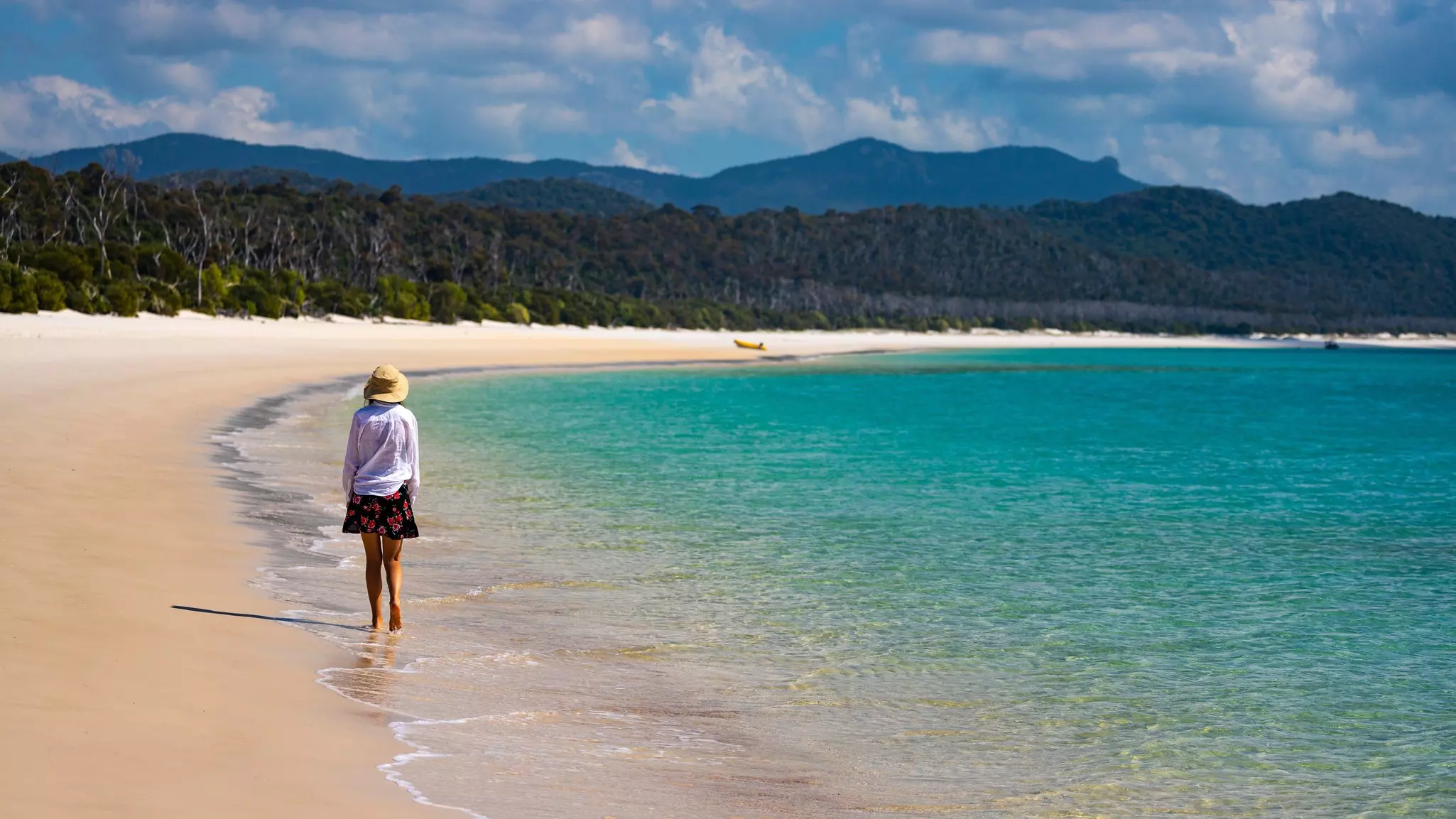 A woman wearing a sunhat, white shirt and skirt walks along a sandy beach by turquoise water in Australia's Whitsunday Islands.