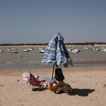 Striped beach umbrella and folding chairs on sand, small boats anchored in the Guadalquivir estuary, Sanlucar de Barrameda.