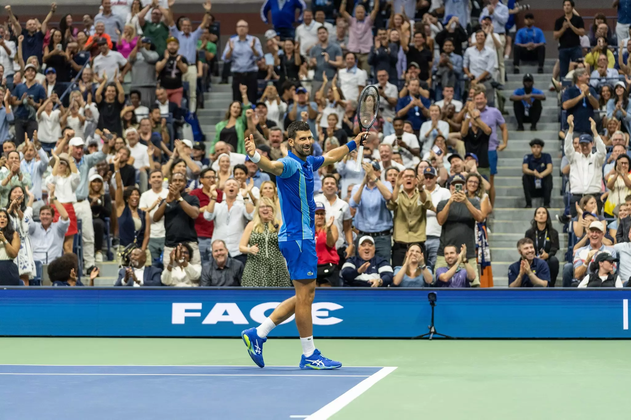 A male tennis player walks off the court with his arms outstrethce smiling as a crowd of people beyind him stand and cheer.