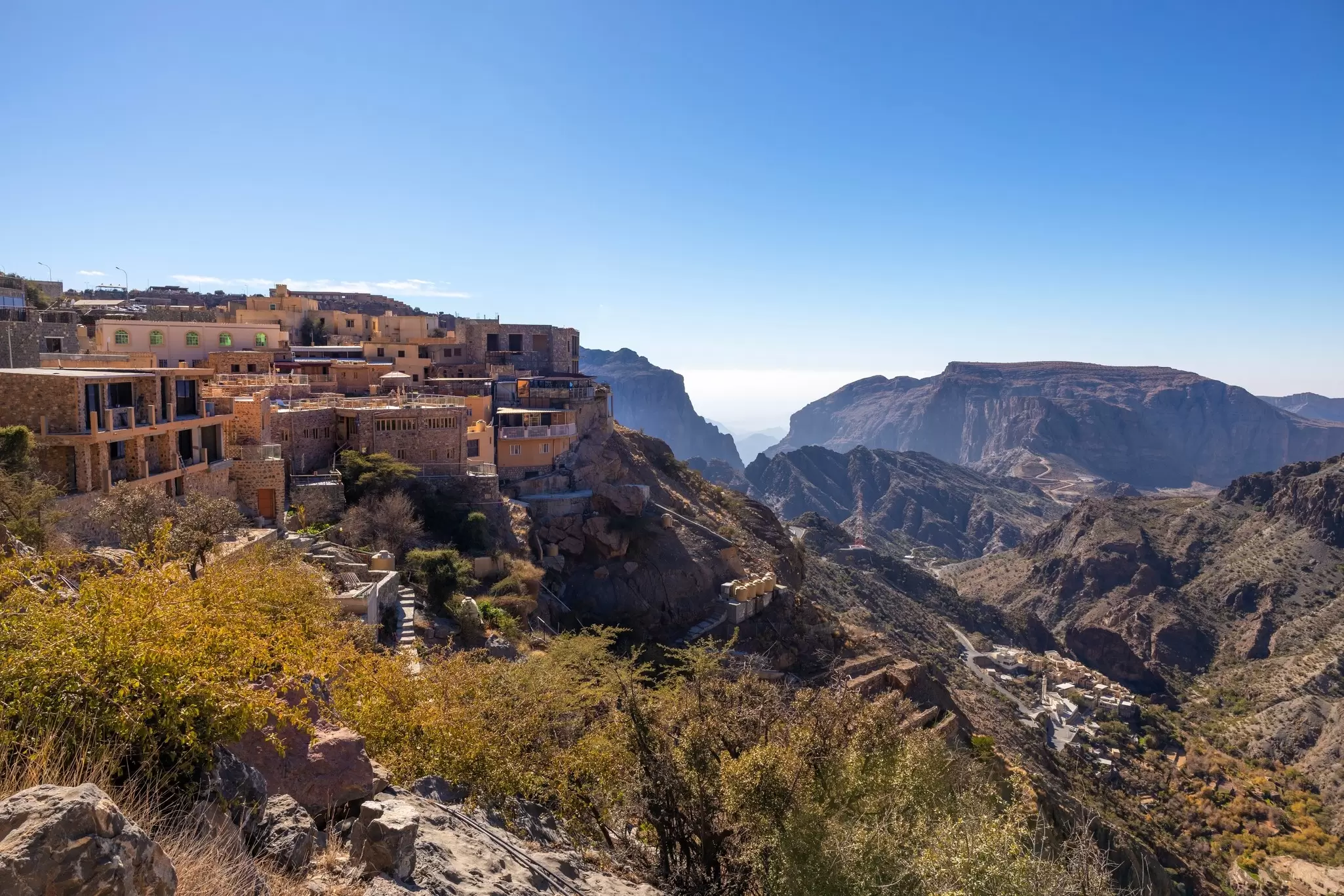 A mountain village bathed in sunlight with a valley of jagged rocks below: Jebel Akhdar, Oman.