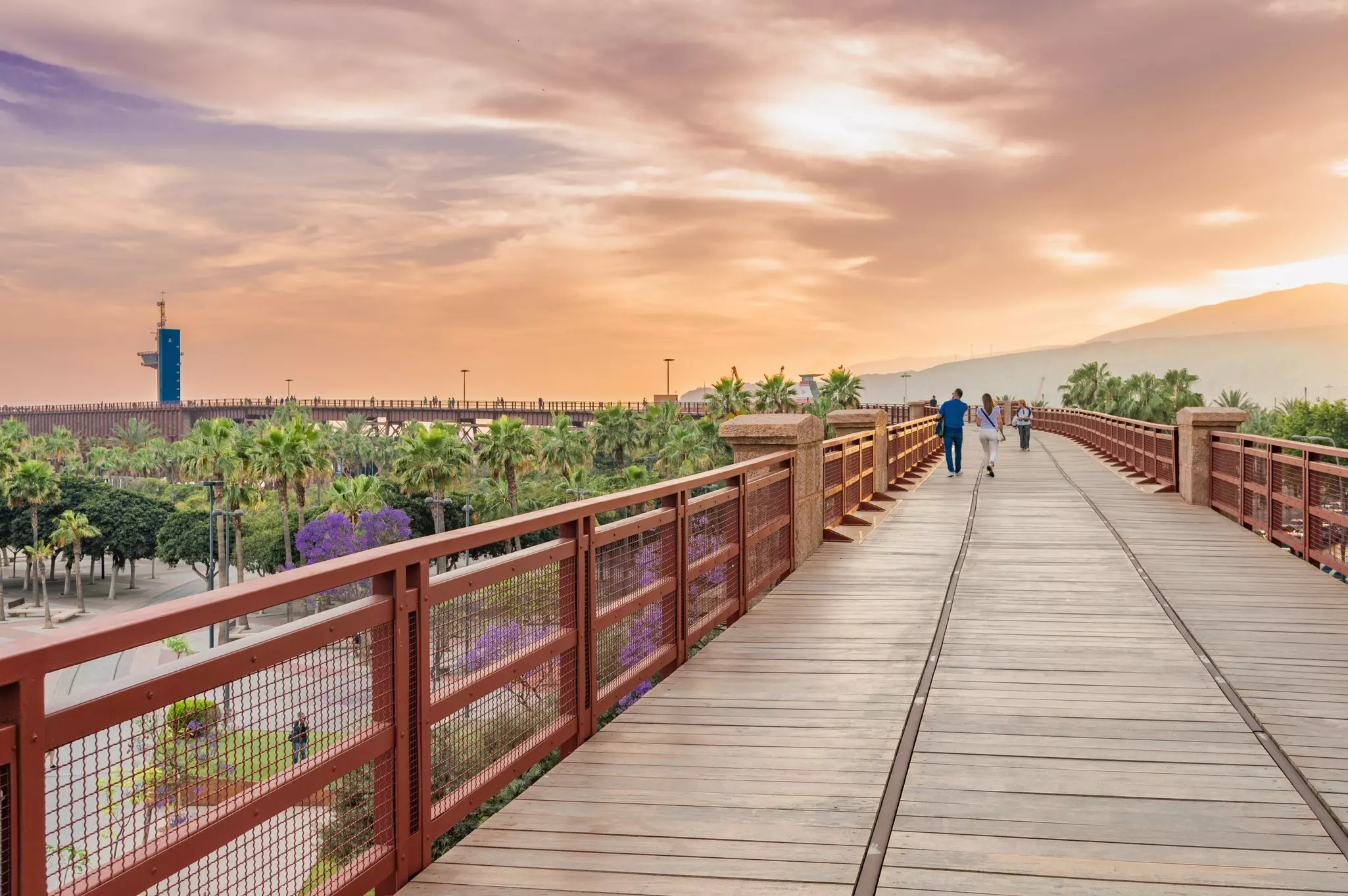 People walk along the top of an industrial structure with old tracks in the center at sunset.
