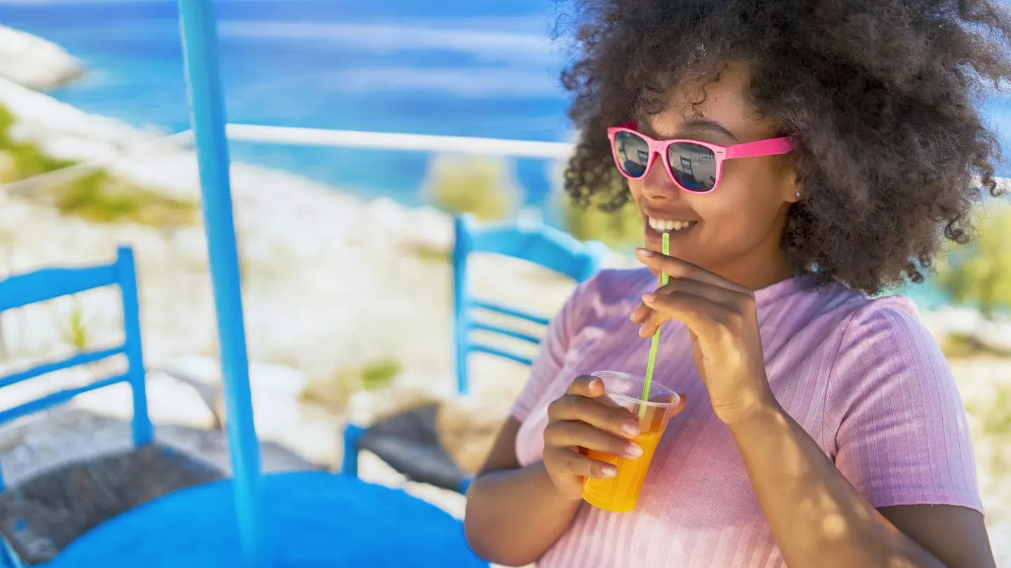 A woman sitting by the sea smiling and drinking a glass of juice