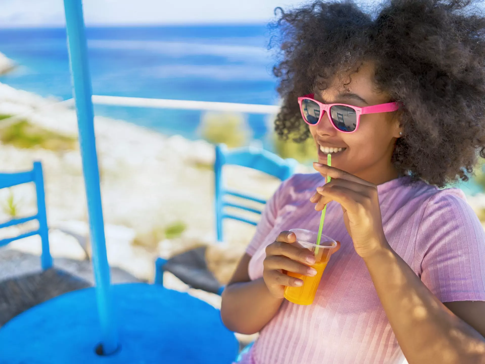 A woman sitting by the sea smiling and drinking a glass of juice