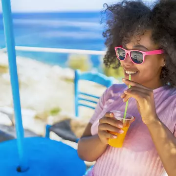 A woman sitting by the sea smiling and drinking a glass of juice