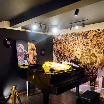 Museum room with a black grand piano draped in a yellow jacket, Freddie Mercury portraits, Queen records and a crowd mural