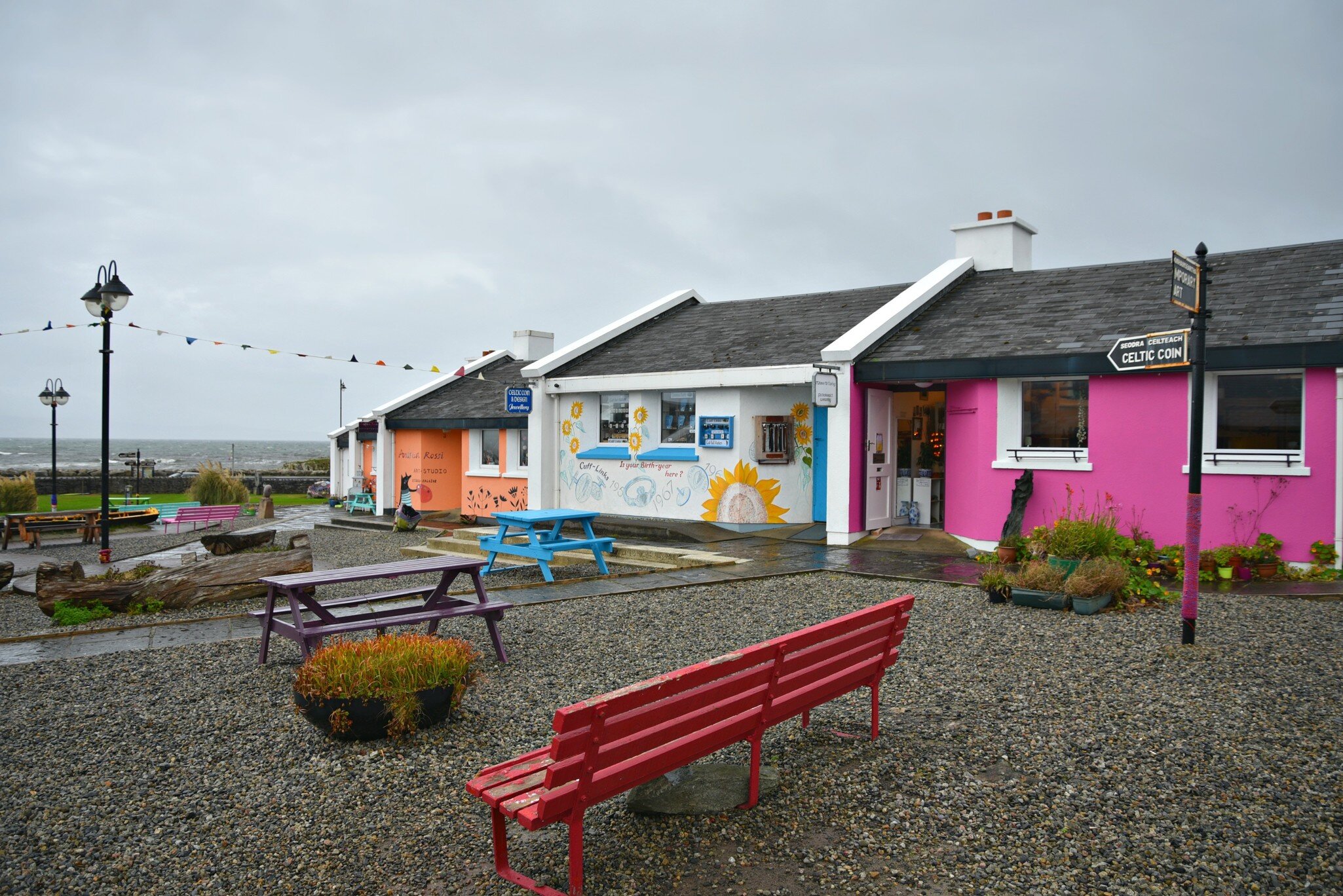 Pink, white and orange cottages with a sunflower mural and 'Celtic Coin' signpost, picnic tables on gravel and the sea beyond