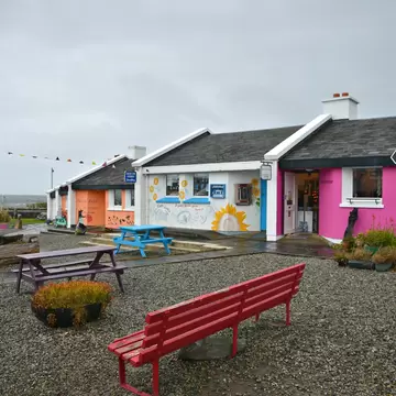 Pink, white and orange cottages with a sunflower mural and 'Celtic Coin' signpost, picnic tables on gravel and the sea beyond